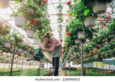 Cheerful young caucasian man using water can for watering the flowers while working at a plant nursery. Young bearded nursery garden worker taking care of the plants in a greenhouse. Copy space. - Powered by Shutterstock