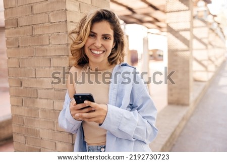 Cheerful young caucasian woman browsing social networks on phone standing on street. Blonde woman with wavy hair wears shirt. Happy weekend concept.