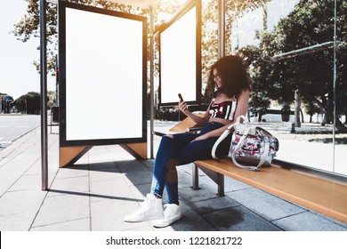 A cheerful young caucasian woman with bulky curly hair is sitting on a yellow metal bench inside of an urban bus stop and using her cell phone, with two white blank info banner templates behind her