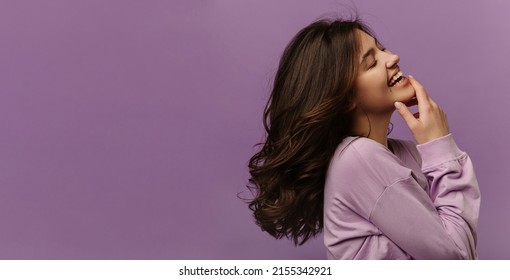 Cheerful young caucasian woman closes eyes and laughs touching her chin on purple background. Dark-haired girl wears casual sweatshirt. Leisure lifestyle and beauty concept - Shutterstock ID 2155342921