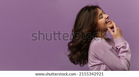 Cheerful young caucasian woman closes eyes and laughs touching her chin on purple background. Dark-haired girl wears casual sweatshirt. Leisure lifestyle and beauty concept