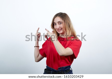 Cheerful young caucasian woman in fashion red shirt looking at camera, pointing fingers behind back on wall isolated on white background in studio. People sincere emotions, lifestyle concept.