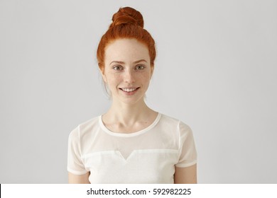 Cheerful Young Caucasian Woman With Freckles And Ginger Hair In Bun Looking And Smiling Happily At Camera, Wearing White Blouse. Redhead Girl With Pretty Joyful Smile Posing At Blank Studio Wall