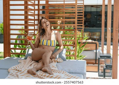 A cheerful young Caucasian woman lounges by the pool, enjoying a refreshing drink on a sunny day, embodying leisure in her casual swimwear. - Powered by Shutterstock