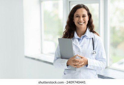 Cheerful young caucasian woman in white medical coat doctor standing by window at clinic hospital, holding laptop computer, online consultation with patient, copy space. Telehealth, remote healthcare - Powered by Shutterstock