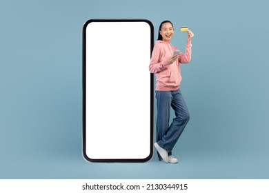 CHeerful Young Chinese Lady With Cell Phone And Credit Card Standing By Huge Mobile Phone With Empty Screen On Blue, Recommending Newest Mobile App For Online Banking, Mockup, Copy Space