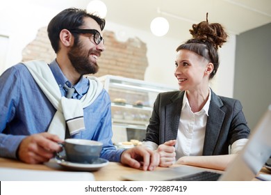 Cheerful young colleagues looking at one another with smiles during conversation at meeting or coffee-break in cafe
