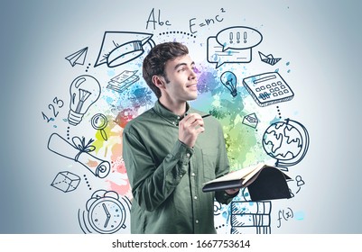 Cheerful Young College Student With Notebook And Pencil Standing Near Gray Wall With Colorful Education Sketch Drawn On It. Concept Of Graduation And Career Choice