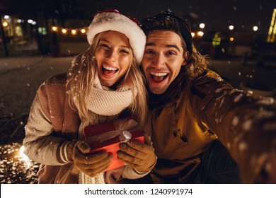 Cheerful young couple dressed in winter clothing holding gift boxes sitting outdoors, taking a selfie, snowfall Stockfoto