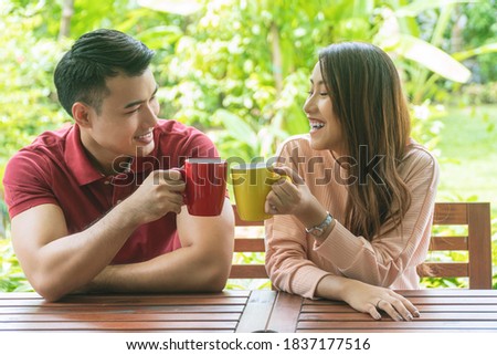 Cheerful young couple holding cup of coffee or tea while sitting on chair in the garden at home. Romantic lover enjoying drink hot mug mocha on the cup glass together in the morning.