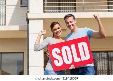 Cheerful Young Couple Holding Sold Sign In Front Of Their Old House