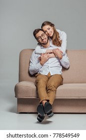 Cheerful Young Couple Hugging On Couch And Smiling At Camera Isolated On Grey