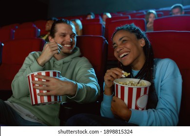 Cheerful young couple laughing, having popcorn while watching movie together, sitting in cinema auditorium. Entertainment and people concept. Horizontal shot