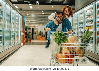 Cheerful young couple laughing and having fun riding a shopping cart filled with groceries in a supermarket, enjoying their time together while shopping for food