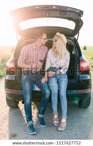 Cheerful young couple looking at mobile phone together while both sitting at the trunk of retro minivan with sea in the background
