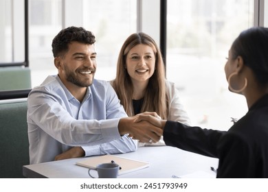 Cheerful young couple shaking hands with African American lawyer, broker, consultant, real estate agent, thanking for consultation. Positive business partners giving handshake at meeting, smiling - Powered by Shutterstock