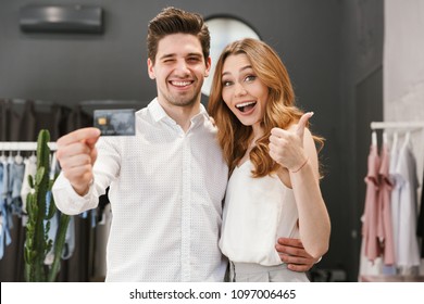 Cheerful Young Couple Shopping For Clothes Together At The Clothing Store, Woman Showing Thumbs Up Gesture And Man Showing Credit Card