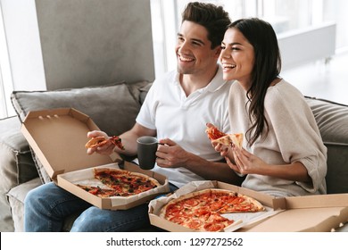 Cheerful Young Couple Sitting On A Couch At Home, Eating Pizza, Watching TV