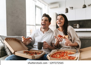Cheerful Young Couple Sitting On A Couch At Home, Eating Pizza, Watching TV