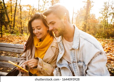 Cheerful Young Couple Sitting On A Bench In The Autumn Park, Using Mobile Phone