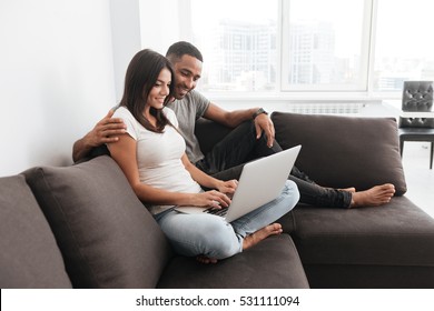 Cheerful Young Couple Smile Together While Using Laptop Indoor. Look At Laptop.