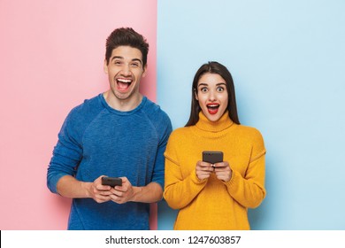 Cheerful young couple standing isolated over two colored background, holding mobile phones