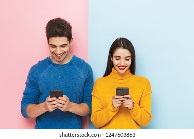 Cheerful young couple standing isolated over two colored background, holding mobile phones