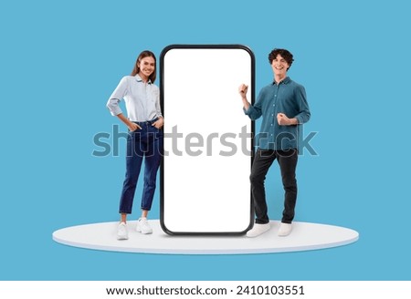 Cheerful young couple stands beside an oversized smartphone, man punching air with joy and woman posing near large blank phone screen, mockup