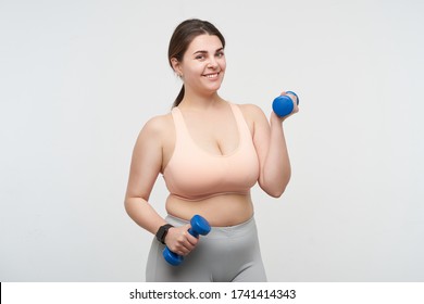 Cheerful Young Cute Brunette Oversized Woman With Chubby Face Smiling Pleasantly At Camera While Training Her Arms With Dumbbells, Isolated Over White Background