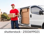 Cheerful young delivery man in red uniform carrying package cardboard box from his white van, delivering parcel to customer