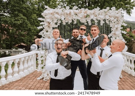 Cheerful, young, energetic witnesses of the bridegroom hold the bridegroom in their arms. Funny photo. The groom in a vest and his friends in white shirts pose near the wedding arch. Wedding in nature