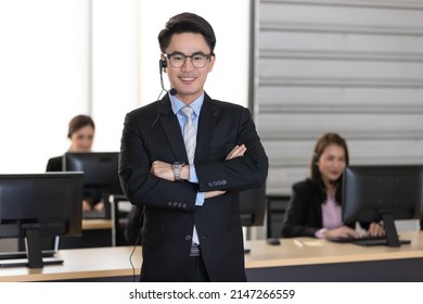 Cheerful Young Ethnic Asian Employee In Elegant Suit And Headset Folding Arms And Looking At Camera In Office With Colleagues Blur In Background. Ready To Support With Good Service Mind Concept.