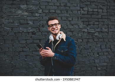 Cheerful young ethnic man in trendy clothes and with headphones surfing internet on mobile phone standing beside brick wall on street and looking at camera