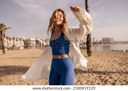 Cheerful young european lady in good mood spends her leisure time on beach near sea. Brown-haired woman wears blue top, shorts and white shirt. Summer vacation concept.