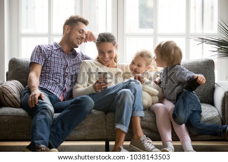 Cheerful young family with kids laughing watching funny video on smartphone sitting on couch together, parents with children enjoying playing games or entertaining using mobile apps on phone at home