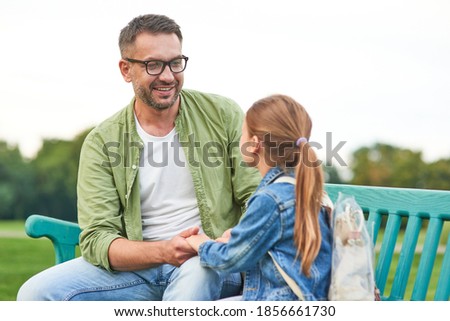Cheerful young father and his cute little daughter talking while sitting on the bench, spending time together in the park on a warm day