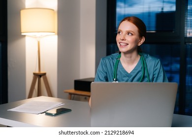 Cheerful Young Female Doctor Wearing Blue Green Medical Uniform Typing On Laptop Computer Looking Away, Sitting At Desk In Hospital Office On Background Of Window At Night.