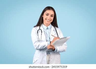 Cheerful young female doctor in white lab coat is writing on clipboard with pen, illustrating attentive patient care and medical record-keeping, blue background - Powered by Shutterstock