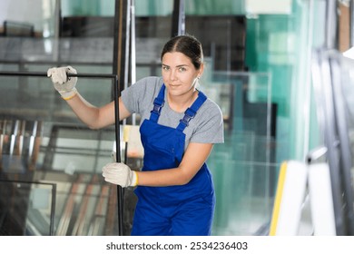 Cheerful young female employee in blue overalls inspecting insulated glass units stacked on storage stands in specialized production workshop.. - Powered by Shutterstock