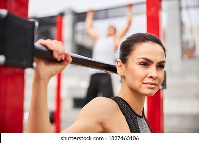 Cheerful young female is exercising with horizontal bar while muscular man is pulling up on sports ground - Powered by Shutterstock