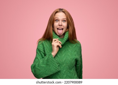 Cheerful young female with long ginger hair touching collar of green sweater and showing tongue while making funny faces against pink background