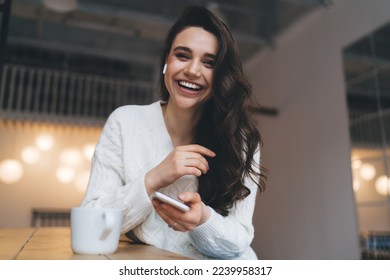 Cheerful young female with long hair in casual clothes browsing mobile phone while sitting at table near coffee cup at home - Powered by Shutterstock