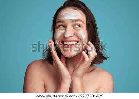 Cheerful young female model with dark hair and bare shoulders smiling happily and looking away wile washing face with foam cleanser against blue background