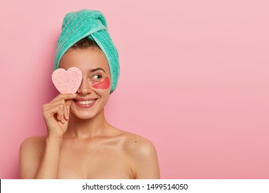 Cheerful young female model has pleasant smile, covers eye with cosmetic sponge, enjoys all benefits of patches, reduces wrinkles, wears wrapped towel on head, has skincare routine after awakening