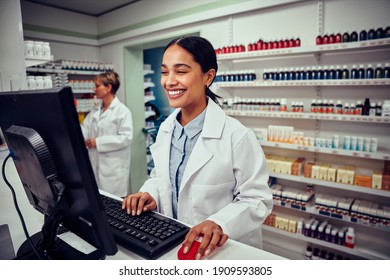 Cheerful young female pharmacist wearing labcoat working on computer in drugstore
