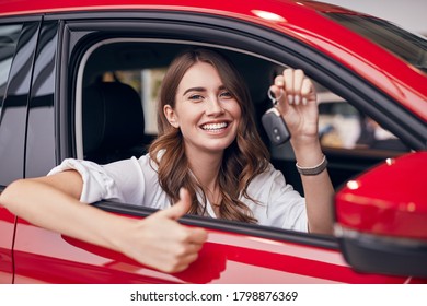 Cheerful young female showing key and thumb up sign while sitting inside new red automobile - Powered by Shutterstock