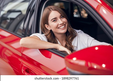 Cheerful young female sitting in shiny red car on passenger seat and looking out open window while enjoying purchase in dealership