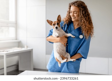 Cheerful young female veterinarian in blue scrubs affectionately cradling Jack Russell Terrier dog during a check-up in a bright veterinary clinic - Powered by Shutterstock