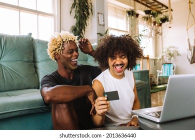 Cheerful Young Gay Couple Smiling Cheerfully While Shopping Online At Home. Two Young Male Lovers Using A Credit Card And A Laptop To Make A Purchase Online. Young Gay Couple Sitting Together Indoors.