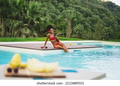 A Cheerful Young Girl Of African Descent Relaxes By Tanning In The Sun With Her Feet In The Pool And Listening To Music With Headphones - Young African American Woman Drinks A Healthy Fruit Cocktail